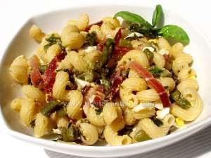 Pasta salad with asparagus and speck, the photo shows a little bowl with short pasta and the ingredients of its sauce: asparagus pieces an tips, speck strips, egg white pieces and aromatic herbs